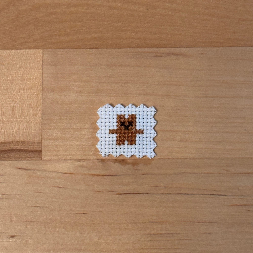 A small cross-stitch sample featuring a brown bear on white fabric, placed on a light wood surface.