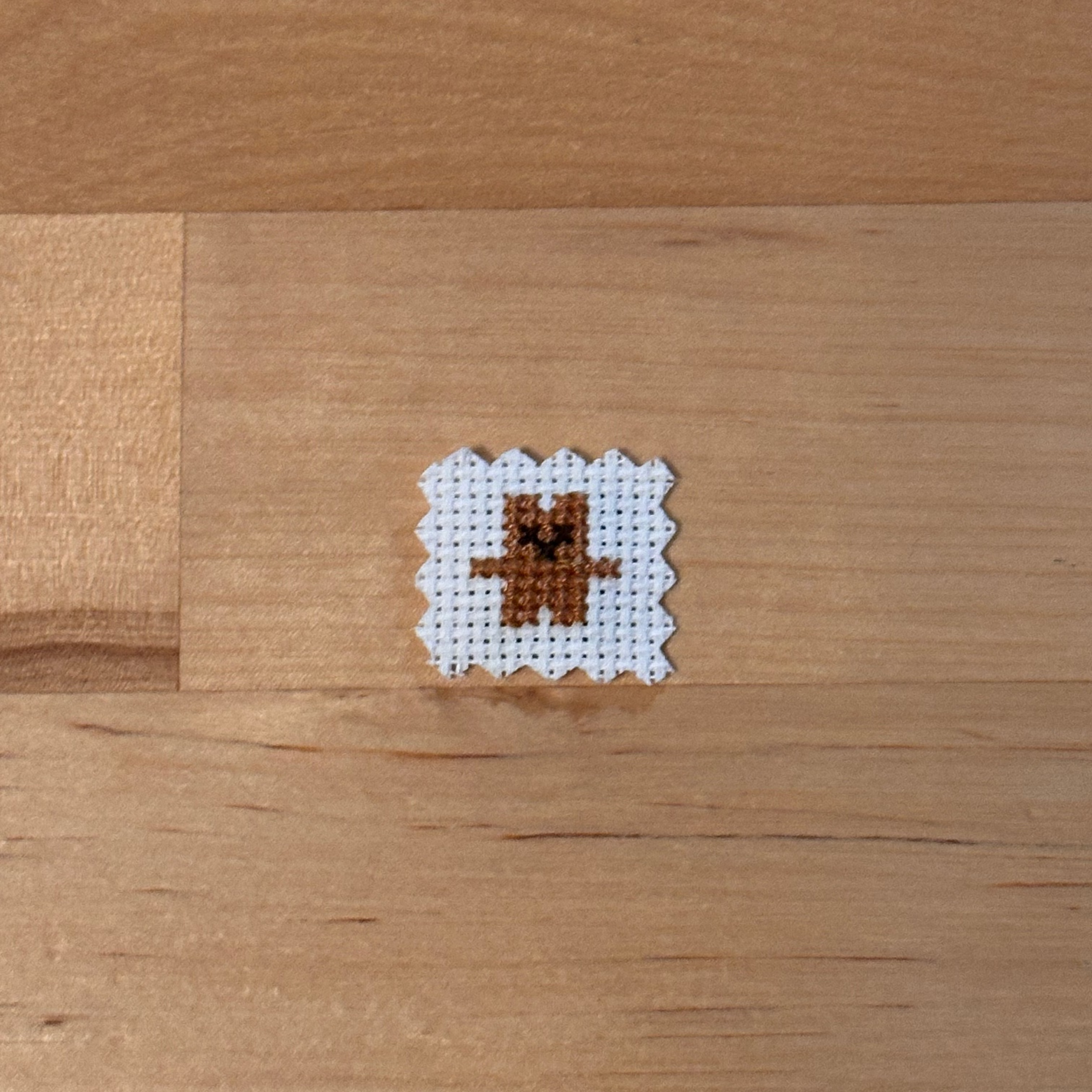 A small cross-stitch sample featuring a brown bear on white fabric, placed on a light wood surface.