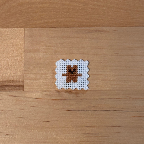 A small cross-stitch sample featuring a brown bear on white fabric, placed on a light wood surface.